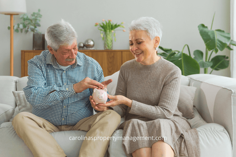 A happy senior couple sitting on a couch, holding a piggy bank and smiling, representing financial savings and smart property decisions with Carolina Property Management. The image highlights the benefits of working with a trusted property management company in Charlotte, NC.