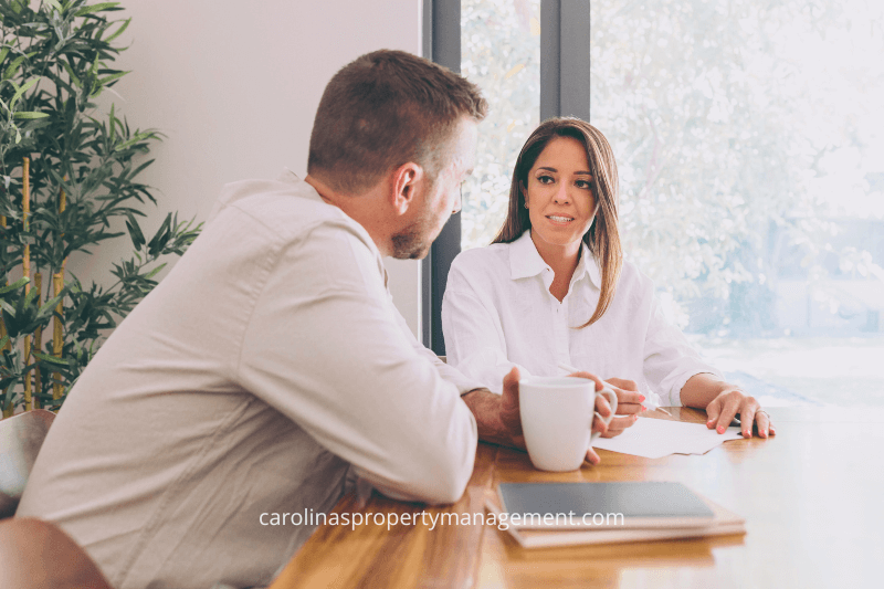 A man and a woman sitting at a wooden table having a discussion, with natural light streaming in from a large window. This image represents a consultation or meeting to discuss rental options, lease terms, or property management services, showcasing the personalized support and expertise offered by Carolinas Property Management Company to help renters and landlords navigate their housing needs effectively.