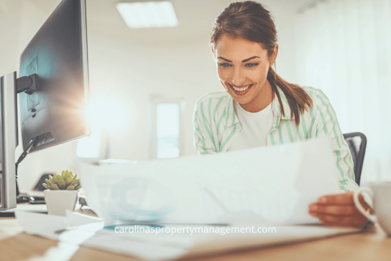 A smiling property manager sitting at a desk, reviewing lease documents with a computer and coffee cup nearby. The image conveys the professional and friendly support provided by Carolinas Property Management Company, ensuring new tenants understand their lease terms and receive thorough guidance when renting an apartment.