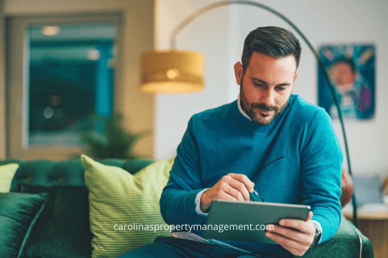 A thoughtful property owner sitting on a green sofa, using a tablet to review tenant applications. This image emphasizes how Carolina Property Management LLC helps property owners by offering a streamlined and reliable tenant screening process compared to Zillow’s application platform.